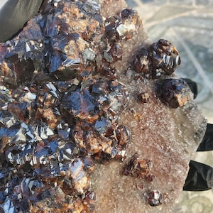 May include: A cluster of dark brown and black crystals on a light brown and white background. The crystals are arranged in a random pattern and have a shiny, metallic finish.