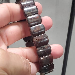 May include: A dark brown bracelet with rectangular beads. The beads have a polished, slightly reflective surface with variations in color. The bracelet is held in a hand, showcasing its design and texture. The bracelet appears to be made of stone or a similar material.