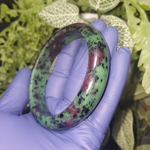 May include: A polished gemstone bangle bracelet. The bracelet features a vibrant mix of green, red, and black colors. The smooth, rounded design of the bracelet is showcased against a background of green foliage.