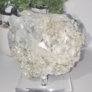 May include: A large, rough-hewn mineral specimen with a mix of textures and colors. The dominant colors are pale green, white, and clear, with crystalline formations. The mineral sits on a clear acrylic stand, showcasing its natural beauty.