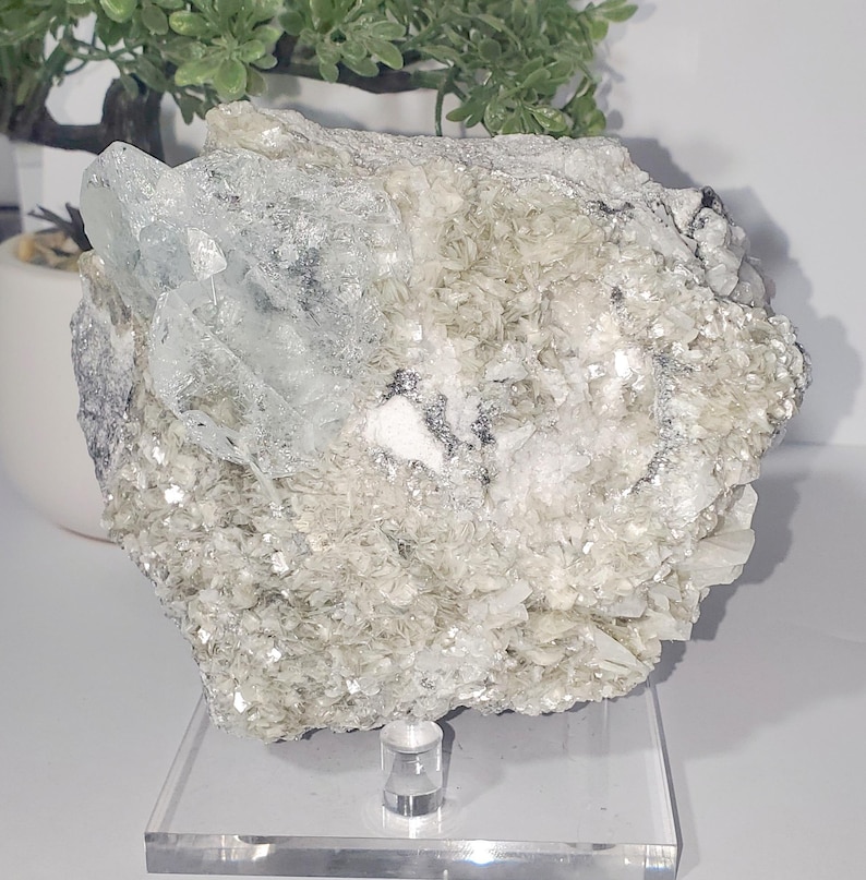 May include: A large, rough-hewn mineral specimen with a mix of textures and colors. The dominant colors are pale green, white, and clear, with crystalline formations. The mineral sits on a clear acrylic stand, showcasing its natural beauty.