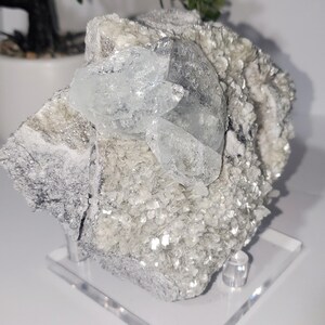 May include: A natural mineral specimen featuring a cluster of clear, light blue crystals atop a matrix of silvery, flaky minerals. The specimen is mounted on a clear acrylic stand, showcasing its natural beauty and texture.