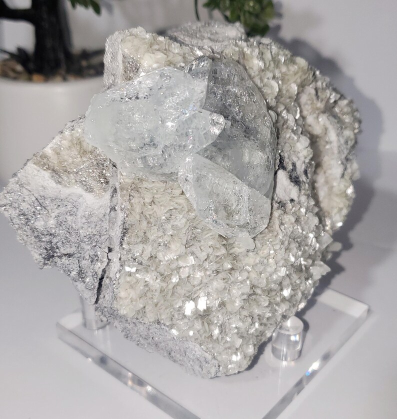 May include: A natural mineral specimen featuring a cluster of clear, light blue crystals atop a matrix of silvery, flaky minerals. The specimen is mounted on a clear acrylic stand, showcasing its natural beauty and texture.