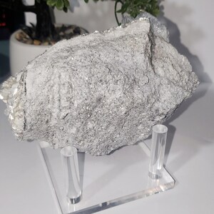 May include: A large, rough-textured, gray and white mineral specimen with sparkling inclusions, displayed on a clear acrylic stand. The mineral has a layered appearance and sits on a rectangular base with two cylindrical supports.