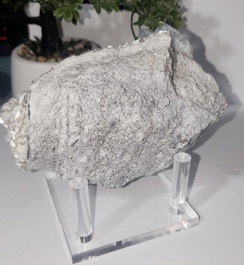 May include: A large, rough-textured, gray and white mineral specimen with sparkling inclusions, displayed on a clear acrylic stand. The mineral has a layered appearance and sits on a rectangular base with two cylindrical supports.