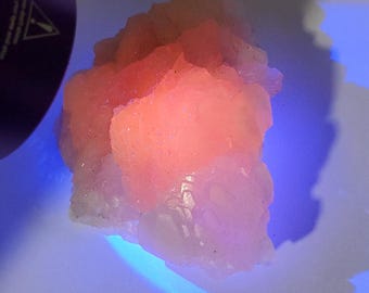 May include: A cluster of pink and white crystals illuminated by a UV light. The crystals have a rough, textured surface and are arranged in a random pattern.