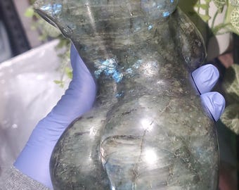 May include: A carved stone torso sculpture in shades of gray and green with blue accents. The sculpture is smooth and polished, with rounded forms suggesting a female figure. The sculpture is held by a person wearing a blue glove.