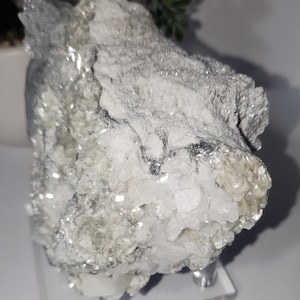 May include: A raw, natural mineral specimen with a crystalline structure. The stone features a mix of white, gray, and silver tones, with a sparkling, reflective surface. The mineral is displayed on a clear acrylic stand.