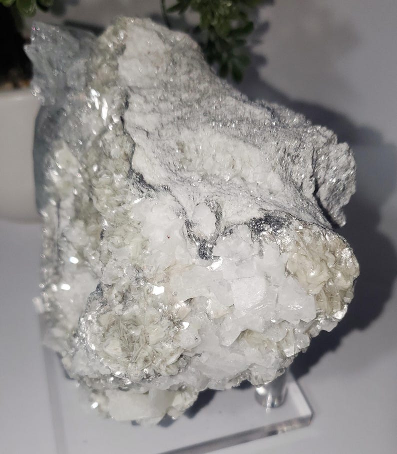 May include: A raw, natural mineral specimen with a crystalline structure. The stone features a mix of white, gray, and silver tones, with a sparkling, reflective surface. The mineral is displayed on a clear acrylic stand.