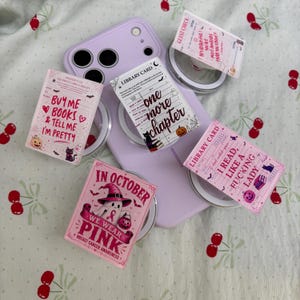 May include: A light purple smartphone case with four rectangular stickers featuring book-themed designs. The stickers display text including "Buy me books & tell me I'm pretty," "one more chapter," "I read like a fucking lady," and "In October we wear pink."