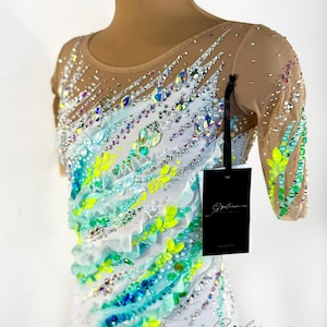 May include: A white leotard with a sheer nude mesh upper, decorated with iridescent rhinestones and colourful accents. The design features turquoise, lime green, and white ruffled details, with a matching headpiece. A black tag hangs from the garment.