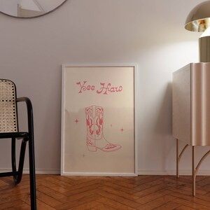 May include: A white framed print with the text "Yee Haw" in pink script. A pink cowboy boot with a white sole is illustrated below the text. The boot has a star design on the shaft and is surrounded by small pink stars.
