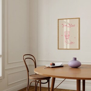 May include: A framed print with a pink cowboy boot illustration and the text "Yee Haw" on a beige background. The print is hanging on a white wall above a wooden table with a vase and a plate of figs.