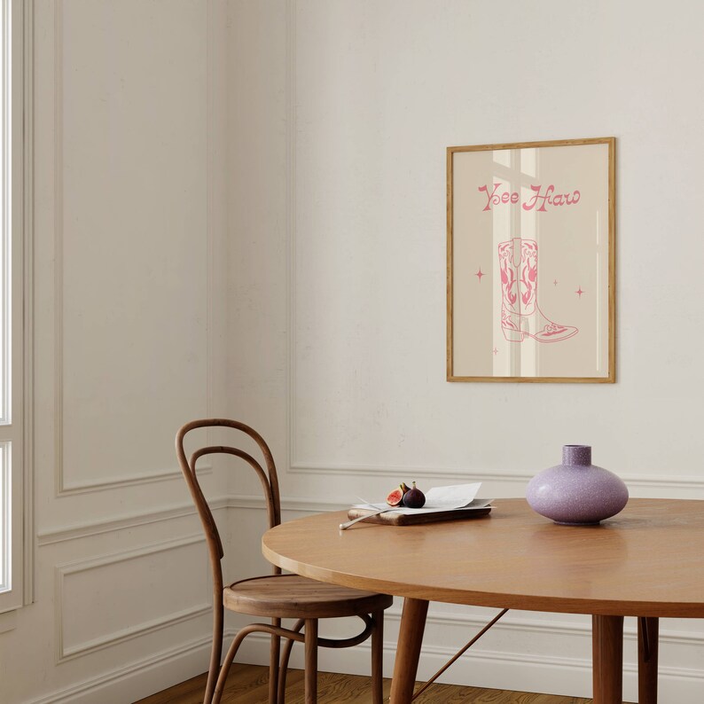 May include: A framed print with a pink cowboy boot illustration and the text "Yee Haw" on a beige background. The print is hanging on a white wall above a wooden table with a vase and a plate of figs.