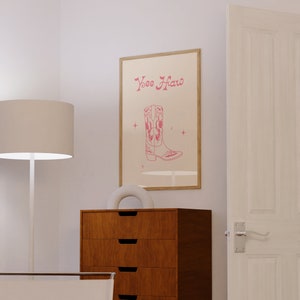 May include: A framed print with a pink cowboy boot and the text "Yee Haw" in pink lettering. The print is hanging on a white wall above a wooden dresser with six drawers.