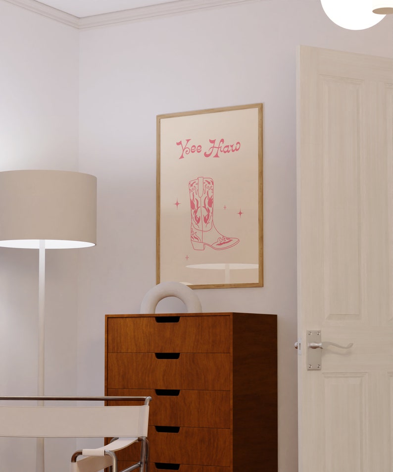 May include: A framed print with a pink cowboy boot and the text "Yee Haw" in pink lettering. The print is hanging on a white wall above a wooden dresser with six drawers.