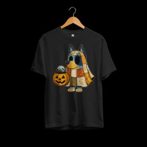 Może przedstawiać: Czarny t-shirt z kreskówkowym psem w kostiumie patchwork trzymającym dynię na Halloween. Pies ma niebieskie i żółte futro oraz patchworkową pelerynę. Dynia jest pomarańczowa z wyrzeźbioną twarzą. Zabawna koszulka z grafiką na Halloween.