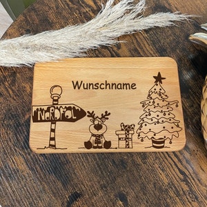 May include: A wooden cutting board with a Christmas theme. It features a reindeer, a Christmas tree, a sign that says "Nordpol", and the text "Wunschname".