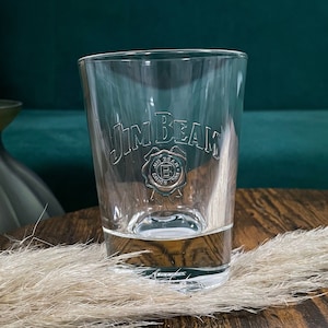 Könnte beinhalten: Ein klares Glas mit der Aufschrift "Jim Beam" auf der Seite. Das Glas hat einen abgerundeten Boden und einen quadratischen Standfuß.