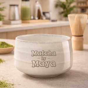 Könnte beinhalten: Eine weiße Keramik-Matcha-Schale mit dem Text "Matcha by Maya". Die Schale steht auf einer hellen Oberfläche, mit einem Bambusbesen und Matcha-Pulver im Hintergrund. Die Schale hat einen Durchmesser von etwa 10 cm.