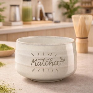Op de afbeelding: Een lichtgekleurde keramische matcha-kom met het woord "Matcha" erop gedrukt. Een bamboe klopper en matcha poeder zijn ook zichtbaar. De kom heeft een licht getextureerd oppervlak.