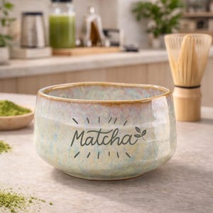 Könnte beinhalten: Eine Keramik-Matcha-Schale mit dem Wort "Matcha" in schwarzer Schrift. Die Schale hat eine hellblaue und beige Glasur. Ein Bambusbesen und ein kleiner Holzlöffel mit Matcha-Pulver sind ebenfalls zu sehen.