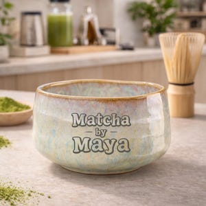 Könnte beinhalten: Eine Keramik-Matcha-Schale mit dem Text "Matcha by Maya" in einem hellblauen und beigen Farbschema. Die Schale ist von Matcha-Pulver, einem Bambusbesen und anderen Matcha-Zubereitungswerkzeugen umgeben.