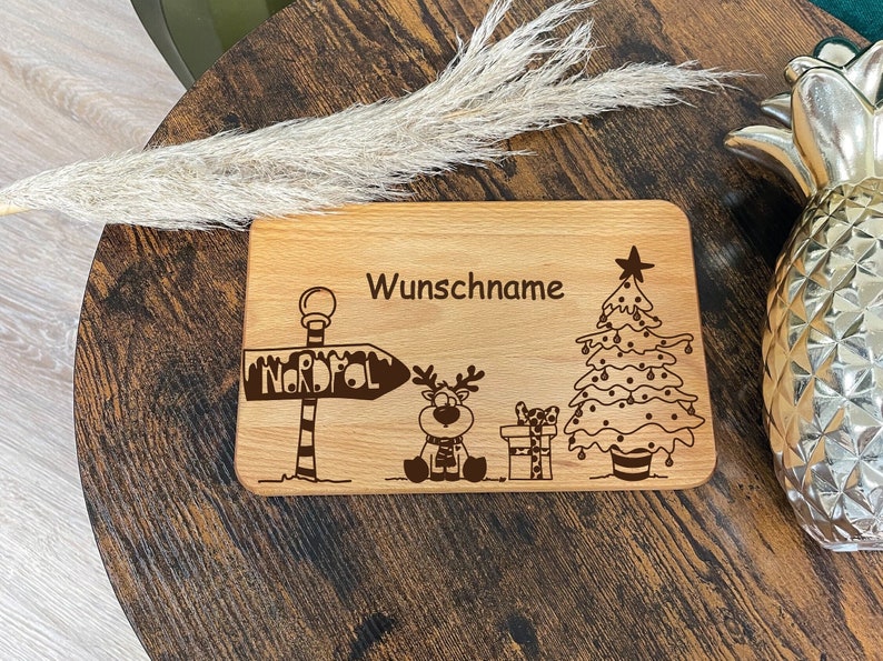 May include: A wooden cutting board with a Christmas theme. It features a reindeer, a Christmas tree, a sign that says "Nordpol", and the text "Wunschname".