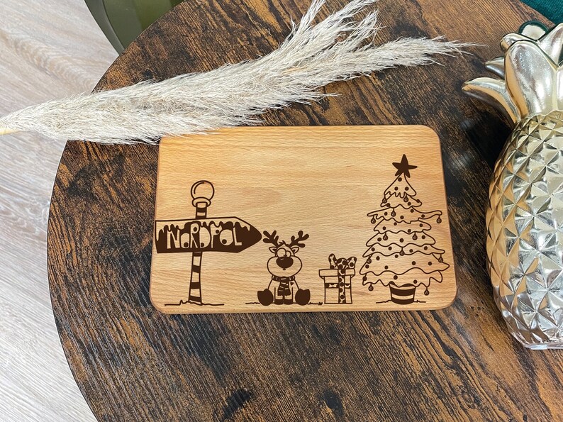 May include: A wooden cutting board with a Christmas theme. It features a reindeer, a Christmas tree, and a sign that says "Nordpol".