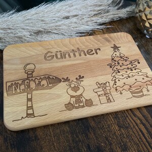 May include: A wooden cutting board with a Christmas theme. It has a reindeer, a Christmas tree, a sign that says "Nordpol" and the name "G&uuml;nther" engraved on it.