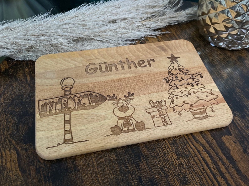 May include: A wooden cutting board with a Christmas theme. It has a reindeer, a Christmas tree, a sign that says "Nordpol" and the name "G&uuml;nther" engraved on it.