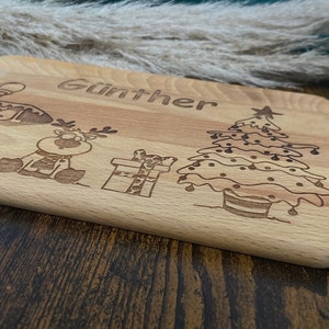 May include: A wooden cutting board with a Christmas theme. It features a reindeer, a Christmas tree, a gift, and the name "G&uuml;nther" engraved on the board.