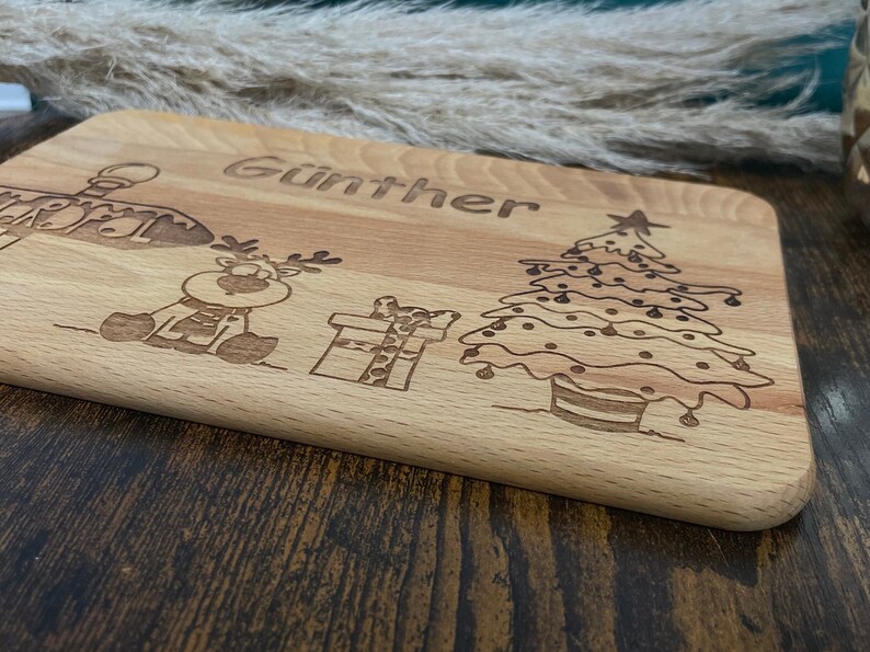 May include: A wooden cutting board with a Christmas theme. It features a reindeer, a Christmas tree, a gift, and the name "G&uuml;nther" engraved on the board.