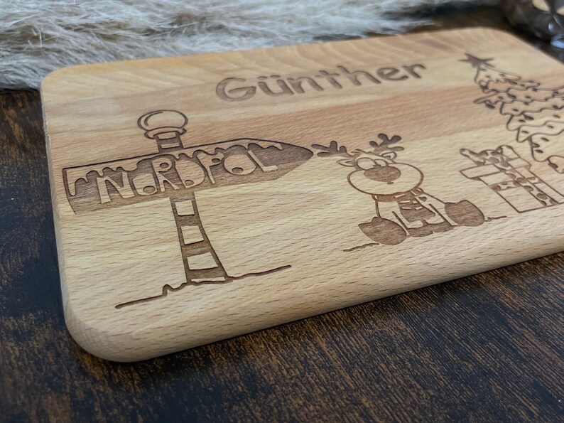 May include: A wooden cutting board with the name "G&uuml;nther" engraved on it. The board also features a cartoon reindeer, a Christmas tree, and a sign that says "Nordpol" (North Pole).