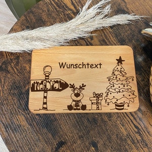 May include: A wooden cutting board with a Christmas theme. It features a reindeer, a Christmas tree, a sign that says "Nordpol" and the text "Wunschtext".
