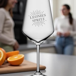Può includere: Un bicchiere da vino trasparente con un lungo stelo, con la scritta "LEONIES SPRITZ" in un carattere stilizzato. Il bicchiere è su una superficie grigia, con fette d'arancia su un tagliere di legno. Per cocktail spritz.