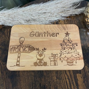 May include: A wooden cutting board with a Christmas theme. It features a reindeer, a Christmas tree, a sign that says "Nordpol", and the name "G&uuml;nther".