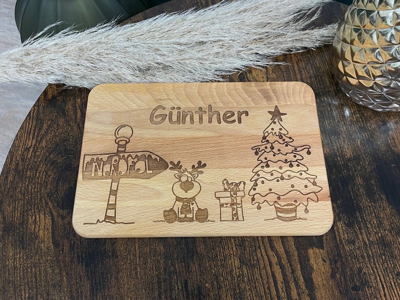 May include: A wooden cutting board with a Christmas theme. It features a reindeer, a Christmas tree, a sign that says "Nordpol", and the name "G&uuml;nther".