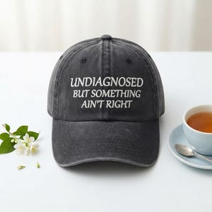 May include: A charcoal grey baseball cap with a distressed, washed finish. The front of the cap features white embroidered text that reads, "UNDIAGNOSED BUT SOMETHING AIN'T RIGHT." The cap has a structured crown, a curved brim, and a button on top.