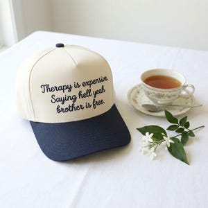 May include: A cream-coloured baseball cap with a dark navy blue brim and a dark navy blue button on top. The front of the cap features the black embroidered text: Therapy is expensive. Saying hell yeah brother is free. The cap has a structured crown.