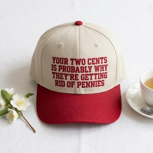 Könnte beinhalten: Eine beige Baseballkappe mit rotem Schirm und rotem gesticktem Text auf der Vorderseite: YOUR TWO CENTS IS PROBABLY WHY THEY'RE GETTING RID OF PENNIES. Die Kappe hat eine strukturierte Krone mit einem Knopf an der Oberseite und Ösen.
