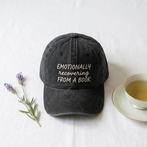 May include: A black baseball cap with a washed-out, vintage texture. The front of the cap features the white embroidered text "EMOTIONALLY recovering FROM A BOOK" in a mix of serif and script fonts. The cap has a curved brim and eyelets.