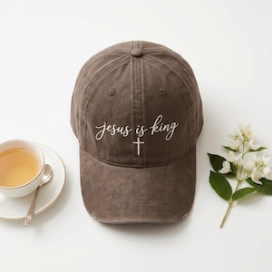Könnte beinhalten: Eine braune Baseballkappe mit dem gestickten weißen Schriftzug "jesus is king" über einem kleinen weißen Kreuzsymbol. Die Kappe hat eine verwaschene, abgenutzte Optik mit einem gebogenen Schirm und einem Knopf oben auf der Krone.