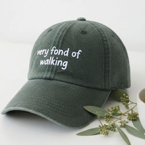 May include: A dark olive green baseball cap with a curved brim and white embroidered text on the front that reads very fond of walking. The cap features a structured crown with stitched eyelets and a fabric strap at the back for adjustment.
