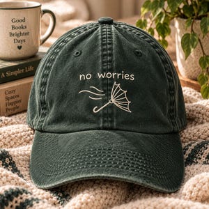 May include: A dark green baseball cap with a washed, distressed finish. The front features white embroidered text that reads "no worries" above a line-art graphic of an umbrella being blown by the wind. The cap has a structured crown and brim.