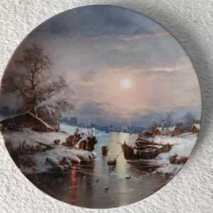 May include: A round ceramic plate with a winter scene painted on it. The scene depicts a frozen river with people ice skating and a village in the background. The sky is a pale blue with a bright sun shining through the clouds.