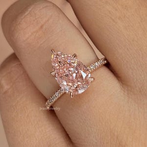 Könnte beinhalten: Ein Ring aus Roségold mit einem großen birnenförmigen, pfirsichfarbenen Mittelstein, der von Krappen gehalten wird, sowie einer dünnen Ringschiene, die mit kleinen weißen Pavé-Diamanten besetzt ist. Der Text JeanJewelry ist auf der Ringschiene sichtbar.