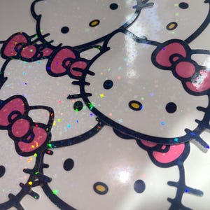 May include: A collection of Hello Kitty stickers. Each sticker features the iconic character's face with a pink bow, black whiskers, and a black outline. The stickers have a white background with a glittery, iridescent finish.