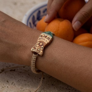 May include: A bracelet featuring a central ceramic fish charm with a textured, metallic gold finish and a teal-colored accent on the tail. The charm is attached to a beige, woven macrame cord band with adjustable pull strings for a secure fit.