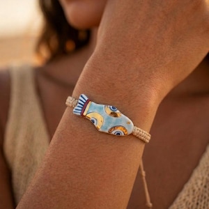 Könnte beinhalten: Ein Armband mit einem Keramik-Fisch-Anhänger in Hellblau, Weiß und Gold mit roten Akzenten, befestigt an einem beigen geflochtenen Band. Der Fisch-Anhänger zeigt ein stilisiertes Augenmuster sowie goldene Details auf der glatten Oberfläche.